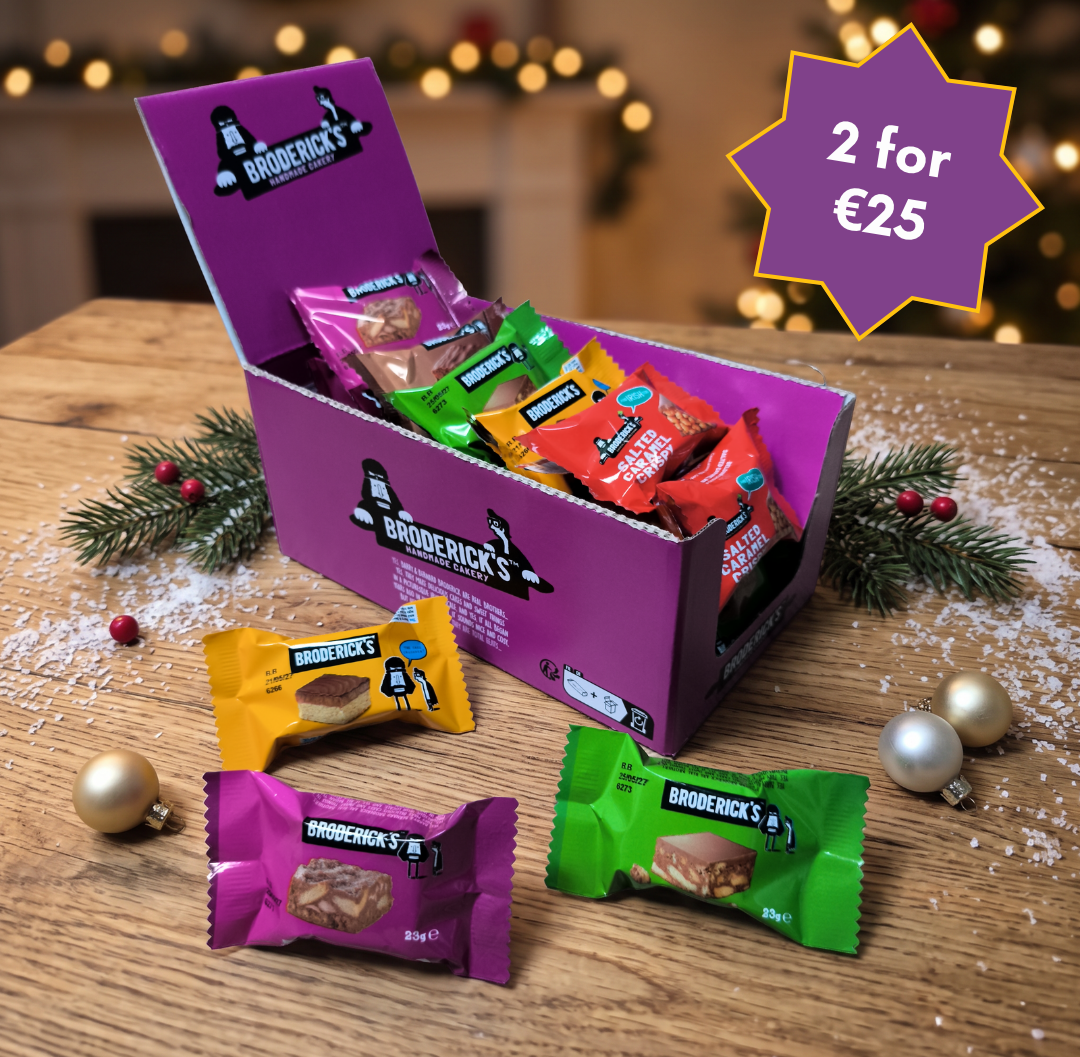 A purple Brodericks chocolate gift box filled with assorted wrapped chocolates sits on a wooden table with holiday decorations, including pine branches, gold ornaments, and blurred Christmas lights in the background. A purple starburst overlay in the top right reads '2 for €25'.